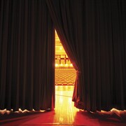 Un rideau de théâtre rouge, entrouvert montre la salle éclairée et des sièges vides.