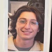 Un jeune homme aux cheveux bruns porte des lunettes et sourit.