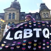 Une personne de dos, debout face à l'édifice de l'Assemblée législative du Nouveau-Brunswick, exhibant un grand drapeau noir décoré de l'acronyme LGBTQ+ en grosses lettres blanches et d'une dizaine de petits coeurs multicolores.