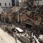 Une rue où plusieurs immeubles sont en ruines, plein de débris étant tombés sur des voitures.