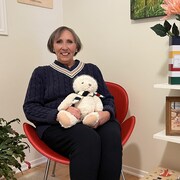 Lise Dumas est assise près d'objets à l'effigie de La Baie, tenant une peluche et portant un pull aux couleurs du magasin.