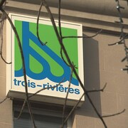 Le logo de la Ville de Trois-Rivières sur un bâtiment avec des branches dénudées de feuilles devant.