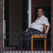 Un homme âgé a l'air inconfortable assis sur une chaise sur un balcon de briques.