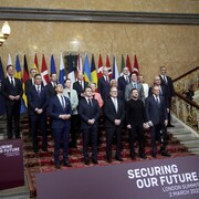 Une vingtaine de personnes posent pour une photo dans un escalier face aux drapeaux représentant les différents pays participant à la rencontre.