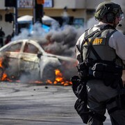 Un policier se tient devant une voiture en flamme.