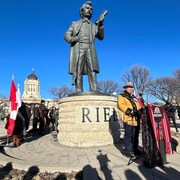 Un homme vêtu d'une veste en suèede perlée parle à un podium. Derrière lui, la statue de Louis Riel et le Palais législatif du Manitoba. 