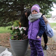 Une fillette vêtue d'un manteau, de pantalons de neige et d'une tuque dépose un bouquet dans une jardinière. Derrière elle, une femme fait de même. 