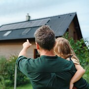 Un père de dos tient sa petite fille dans ses bras et lui pointe les panneaux solaires sur le toit d'une maison.