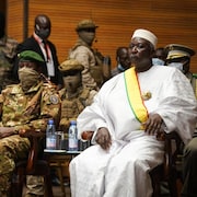 Le président, vêtu en blanc, est assis à côté du vice-président, en treillis militaire. Plusieurs militaires en uniforme sont derrière eux. 