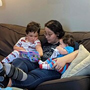 Une maman est assise sur un divan entourée de ses deux jeunes enfants. Ils regardent un livre. 