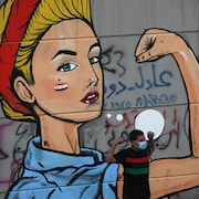 Un homme en face d'une murale montrant une femme blonde avec un bras levé, les manches retroussées.