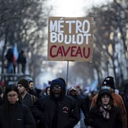 Des personnes rassemblées dans une rue. L'une d'entre elles tient une pancarte sur laquelle est écrit : « Métro, Boulot, Caveau ».