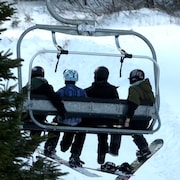 Quatre personnes chaussées de planches à neige sont assises sur un télésiège.
