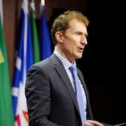 Marc Miller en conférence de presse à Ottawa, devant les drapeaux du Canada et des provinces et territoires.
