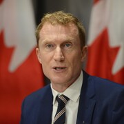 Marc Miller en conférence de presse devant deux drapeaux du Canada.