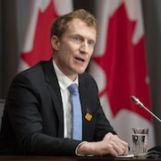 Marc Miller en conférence de presse devant plusieurs drapeaux du Canada.
