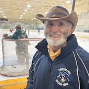 Un homme avec un chapeau de cow-boy derrière la baie vitrée en arrière du gardien de but dans un aréna pendant une pratique de hockey.