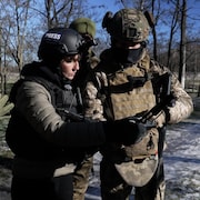 La journaliste, v&ecirc;tue d'un casque et d'un gilet pare-balles, est entour&eacute;e de trois soldats. Ils sont dans un espace bois&eacute;. La journaliste tient un d&eacute;tecteur de drones, elle regarde son &eacute;cran.