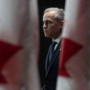 Mark Carney, de profil, entre deux drapeaux du Canada.