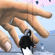 Un passant masqué marchant devant une main gigantesque sur une fresque murale.