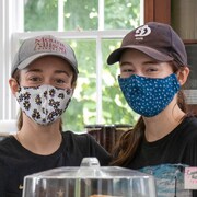 Deux jeunes femmes coiffées de casquettes et le visage couvert d'un masque de tissu sont debout derrière le comptoir du café où elles travaillent.