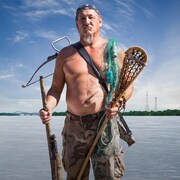 L'homme, torse nu, debout devant le fleuve Saint-Laurent, porte un arc, une carabine, un bâton de crosse et a un filet de pêche sur l'épaule.