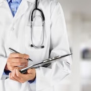 Un médecin dans un couloir d'hôpital s'apprête à écrire sur un document. Il porte un stéthoscope autour du cou.