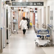 Une femme vêtue d'un sarrau blanc marche dans un corridor d'hôpital où l'on voit une civière. 