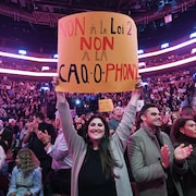 Une femme souriante tient au bout de ses bras une pancarte qui porte le message &laquo; Non &agrave; la loi 2, non &agrave; la caq-o-phonie &raquo; au milieu de la foule qui remplit le Centre Bell.