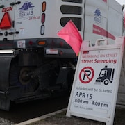 Une pancarte de la Ville de Calgary qui indique une interdiction de stationnement pour déblayage des rues à partir du 15 avril de 8 h à 16 h sous peine de recevoir une amende. Derrière, un des camions de nettoyage.