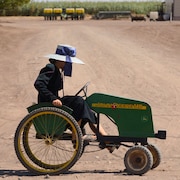 Une femme portant une robe traditionnelle mennonite et un chapeau gris au ruban bleu pédale sur un quadricycle vert de marque John Deere, sur une route de terre et de sable.