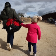 Une femme et deux fillettes de dos marchent à côté du mur frontalier.