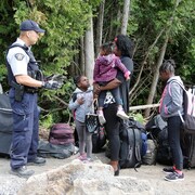 Une femme et ses trois enfants entourés de valises parlent à un agent armé.