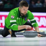 Le capitaine de l'équipe de la Saskatchewan, Mike McEwen, vêtu de vert, s'apprête à envoyer une pierre en s'appuyant sur son balai.