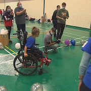 Une jeune fille se déplace en fauteuil roulant dans un gymnase. Elle est entouré de gens qui l'encouragent. 
