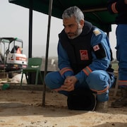 Un homme aux cheveux gris est accroupi pr&egrave;s d'une tombe. Il est en habit de secouriste bleu, un casque est &agrave; ses pieds. Il porte un gilet pare-balles.