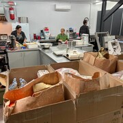 Des personnes préparent des repas dans une cuisine. Des boîtes d'aide alimentaire se trouve à l'avant.