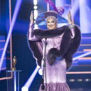 Une drag queen vêtue de mauve sourit. 