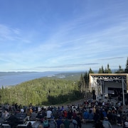 Spectacteurs assis dans un ampithéatre naturel, devant une scène avec au loin une vue sur la forêt, la baie, les rivières et le fleuve. 