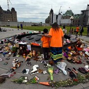 Une grand-mère et son petit-fils, de dos, se recueillent près de la Flamme du centenaire à Ottawa, avec des dizaines de paires de souliers et de fleurs.