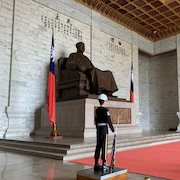 Un garde de sécurité devant une grande statue représentant un homme en robe, assis.