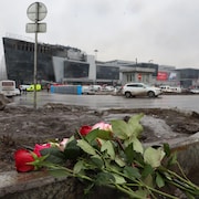 Un bouquet de rose déposé sur un plot de béton, avec de la boue derrière, une route, un bâtiment incendié et un ciel gris.