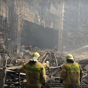 Un immeuble en ruine entouré de fumée et deux hommes en tenue jaune, casqués, qui regardent les dégâts. 