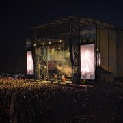 Le groupe Mumford and Sons joue une chanson sur la grande scène de Cavendish. Il fait nuit. La lumière de la scène éclaire la foule.