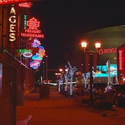 Un plan plus large de la rue, avec des signes néons qui brillent des deux côtés de la route. Au fond, la silhouette de la place Rogers se dessine.