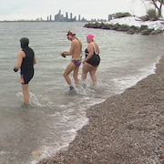 Trois personnes en costumes de bain, les pieds dans l'eau alors que la neige recouvre  une rive proche.