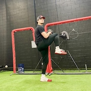 Un homme portant une casquette est sur le point de lancer une balle de baseball dans un centre intérieur, sur un gazon synthétique.