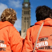 Deux personnes portant un chandail avec le slogan «Chaque enfant compte» posent de dos devant le parlement à Ottawa.