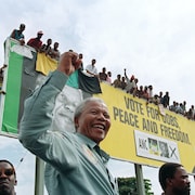 Souriant et le poing levé, Nelson Mandela est debout devant un panneau sur lequel se tiennent de nombreuses personnes.