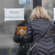 Une femme de dos, qui s'apprête à pousser la porte d'entrée vitrée d'un édifice. Sur la porte sont collées deux affiches qui disent : « Nous embauchons ».